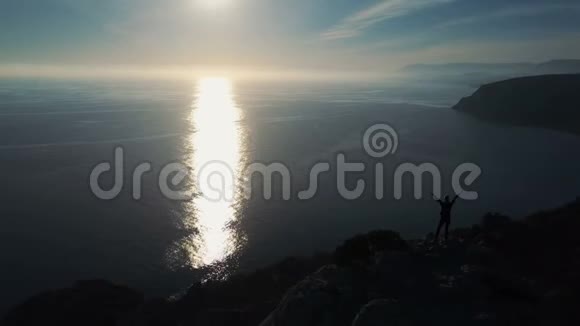 日出时年轻女子站在山顶上靠海的空中轮廓快乐的年轻女子视频的预览图