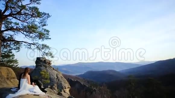 美丽迷人的幸福新娘和时尚的新郎坐在阳光明媚的迷人山脉的背景上视频的预览图