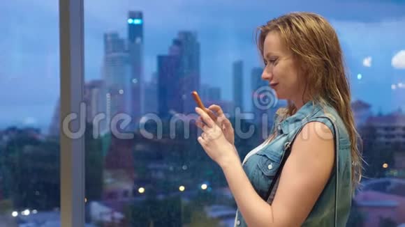 站在摩天大楼背景下窗前的湿漉漉的金发女孩在外面下雨的时候用手机视频的预览图