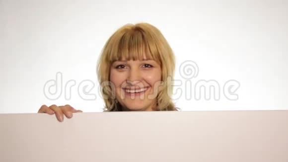 微笑的幸福女人站在后面靠在一个白色的空白广告牌或标语牌上拇指指向顶部躲藏起来视频的预览图