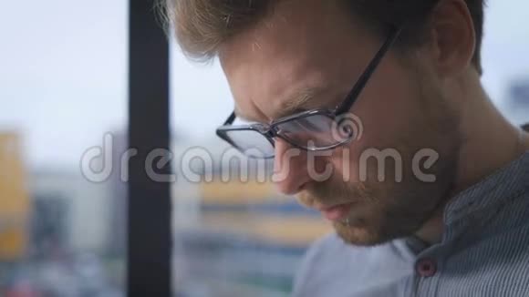 戴眼镜的年轻人坐在大厅的窗台上一边看着平板电脑上的新闻视频的预览图