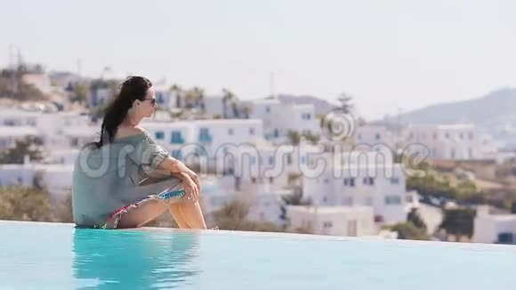 美丽的年轻女孩在室外游泳池边缘放松美丽的Mykonos景色视频的预览图