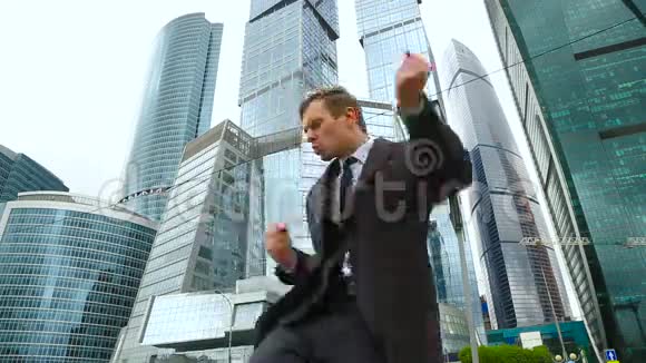 快乐的商人在一座商业中心摩天大楼的背景下在一条城市街道上跳舞视频的预览图