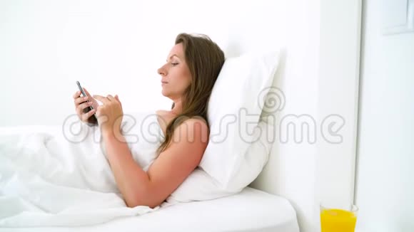 可爱的女人早上检查智能手机在床上喝橙汁视频的预览图