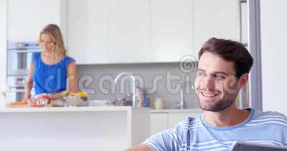 微笑的男人用平板电脑和他的妻子在他身后准备饭菜视频的预览图