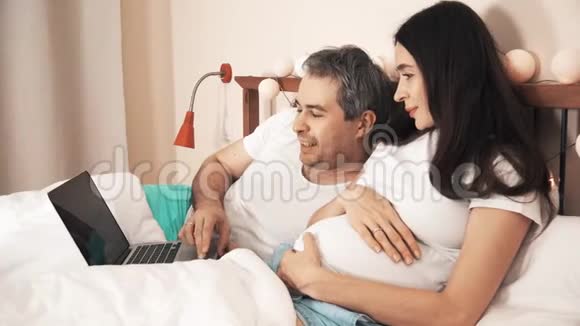 丈夫和怀孕的妻子在床上用笔记本电脑视频的预览图