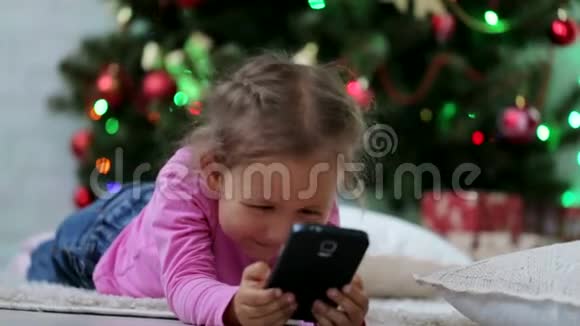 小可爱的女孩用智能手机在圣诞树前特写镜头视频的预览图