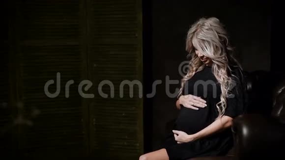 年轻的孕妇穿着黑色礼服坐在前面在黑暗中慈爱地抚摸着她的腹部视频的预览图