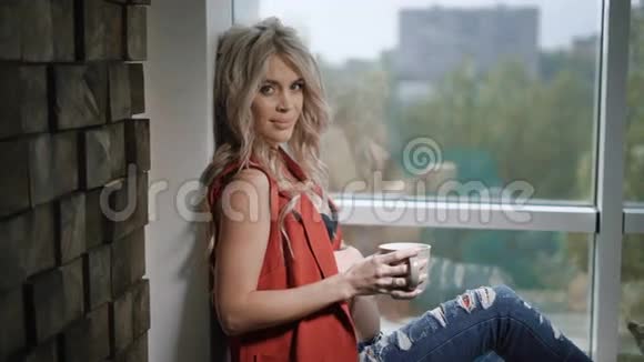 穿着红色衬衫和蓝色牛仔裤的美丽怀孕金发女人坐在靠近窗户和墙壁的窗台上视频的预览图