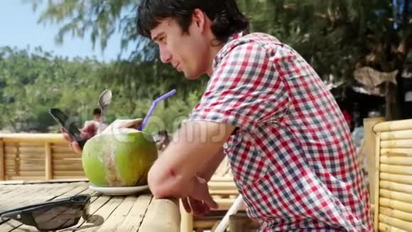 英俊的年轻商人戴着太阳镜在海滩咖啡馆喝着新鲜的椰子汁在棕榈树上的海景视频的预览图