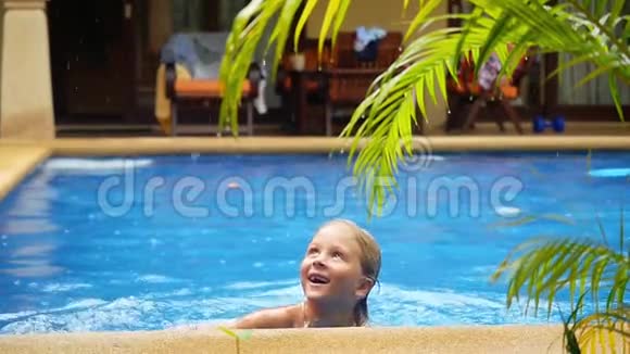 快乐的男孩在雨中用一棵棕榈树的树枝在一个有蓝色水的游泳池里游泳慢动作1920x1080视频的预览图