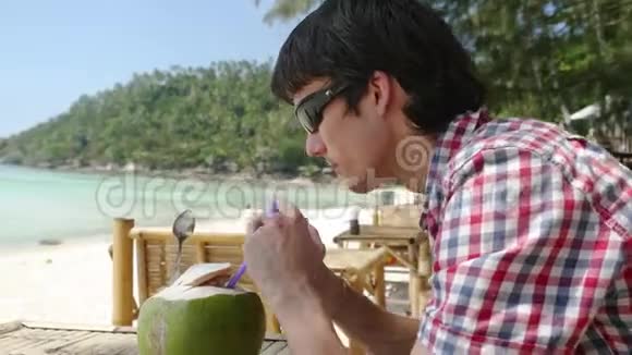 英俊的年轻商人戴着太阳镜在海滩咖啡馆喝着新鲜的椰子汁在棕榈树上的海景视频的预览图