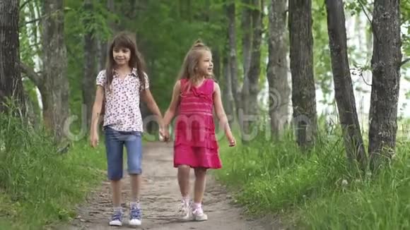两个小女孩牵着手走在绿色的小巷里孩子们在户外散步两个小妹妹慢动作视频的预览图