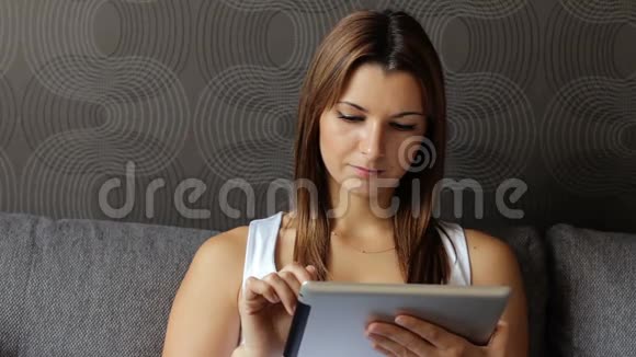 年轻微笑的女人看数字平板电脑的肖像有平板电脑的女人坐在沙发上女人用平板电脑视频的预览图