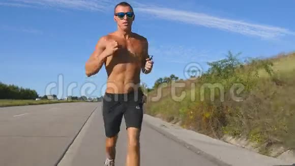 年轻强壮的肌肉男戴着太阳镜在高速公路上慢跑用他的智能手机耳机听音乐男性视频的预览图