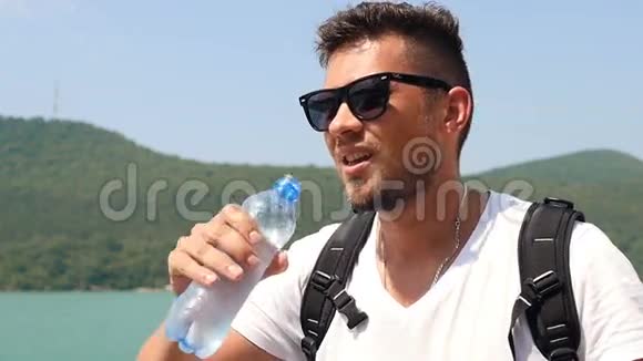 阳光明媚的夏日太阳玻璃的年轻人在山湖的背景下喝着纯净的水视频的预览图