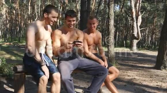 三个快乐的肌肉运动员坐在森林里看着智能手机的照片快乐的朋友视频的预览图