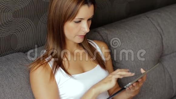 年轻微笑的女人看数字平板电脑的肖像有平板电脑的女人坐在沙发上女人用平板电脑视频的预览图