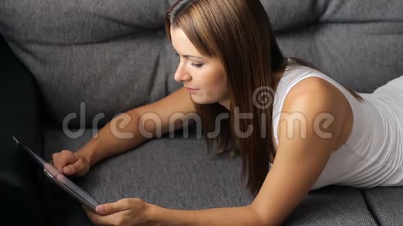 女人躺在沙发上用平板电脑年轻微笑的女人看数字平板电脑的肖像在视频的预览图