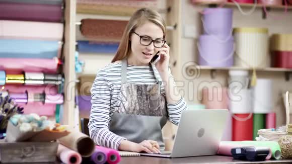 花店女花店女花匠在手机上聊天的同时使用笔记本电脑在线查看订单批发花卉销售视频的预览图