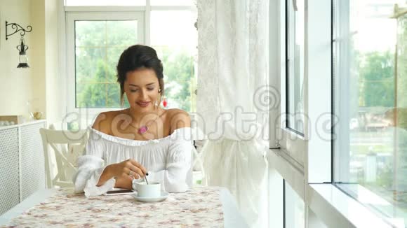 一个穿着白色衬衫的深褐色美女在明亮的露台上喝咖啡视频的预览图