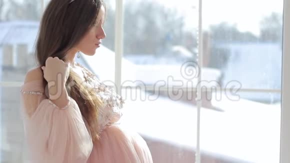 怀孕的女孩坐在窗户附近穿着一件漂亮的粉红色连衣裙视频的预览图
