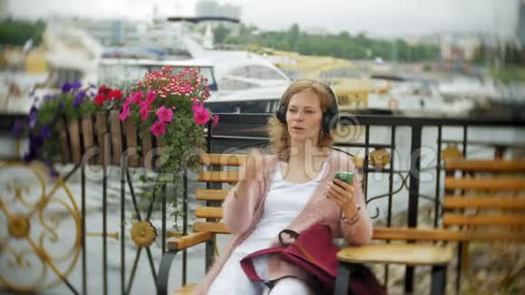 一个女孩用智能手机坐在沙滩上的花凳上听着耳机里的音乐跳舞游艇和视频的预览图