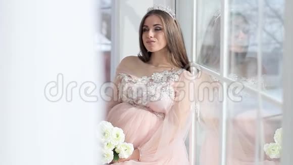 怀孕的女孩坐在窗户附近穿着一件漂亮的粉红色连衣裙视频的预览图