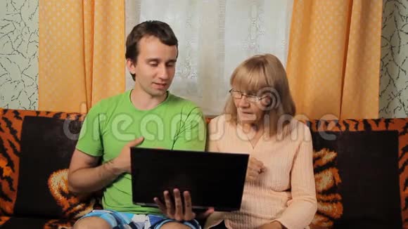 成年男性教为笔记本电脑工作的老妇人母子俩坐在家里的沙发上视频的预览图