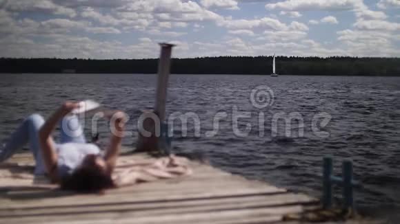 年轻女子躺在模糊的木墩上看书帆船在背景集中慢动作视频的预览图