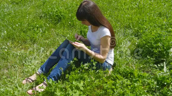 年轻的白种人黑发女孩坐在草地上的草地上在公园里使用数字平板电脑快乐的女人视频的预览图