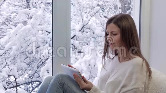 漂亮的女孩坐在窗台上看着窗户用平板电脑冬天在外面视频的预览图