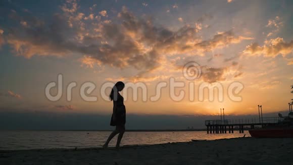 日落时海滩上一个穿着浅色连衣裙的女孩的剪影美丽的少女赤脚走在沙滩上慢动作视频的预览图