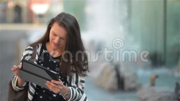 快乐的年轻女子带着平板电脑坐在街边的长椅上在喷泉背景下进行视频聊天视频的预览图