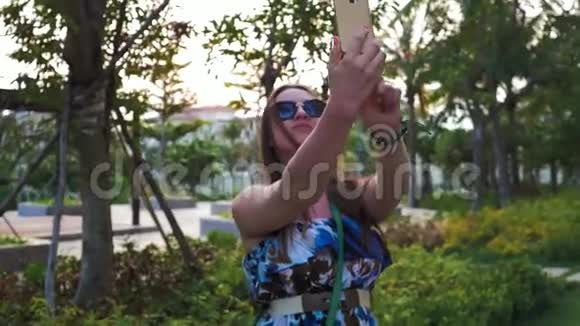 漂亮的快乐女孩戴着眼镜和蓝色连衣裙在手机上摆出自拍绿色的手掌和美丽的阳光中等视频的预览图