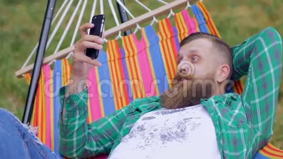 帅哥躺在吊床上在户外使用智能手机视频的预览图