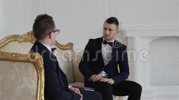 两位穿着蝴蝶结西装的商人正在一间漂亮的白色房间里与优雅的金色沙发交流视频的预览图