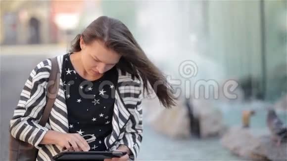 美丽的女孩阅读和浏览电子书或平板电脑坐在长凳上的街道喷泉背景视频的预览图