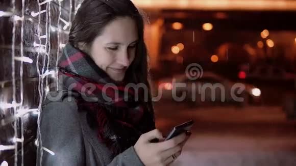 年轻迷人的女人在圣诞夜的大雪中使用智能手机站在灯光墙附近视频的预览图