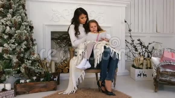 年轻的母亲和她的小女儿在壁炉旁的扶手椅上轻松地读着圣诞故事视频的预览图