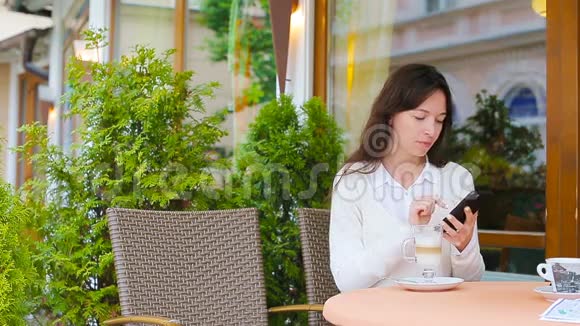 在户外咖啡馆使用智能手机的年轻女孩散步后的美女坐在露天咖啡厅喝咖啡视频的预览图