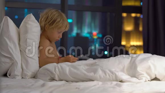 晚上坐在床上的小男孩玩平板电脑背景是一座摩天大楼的轮廓移动视频的预览图