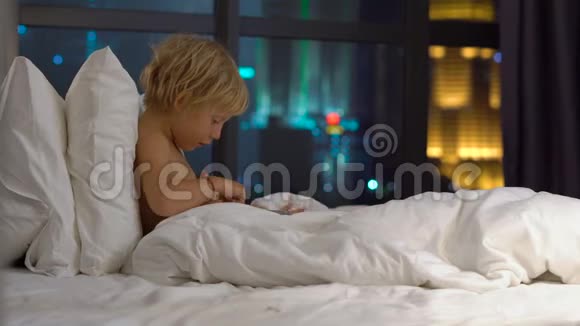 晚上坐在床上的小男孩玩平板电脑背景是一座摩天大楼的轮廓移动视频的预览图
