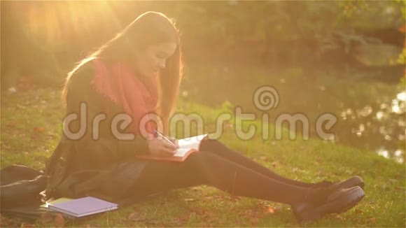 美丽的学生女孩坐在草地上在美丽的公园里写着笔记本湖在背景上视频的预览图