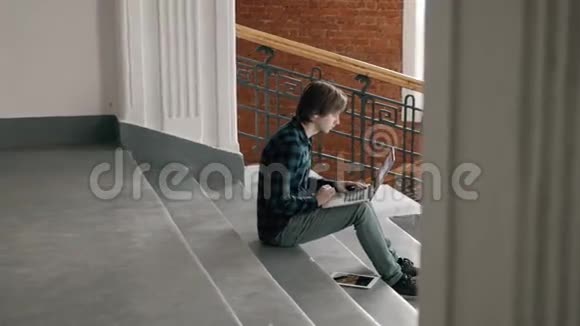 自由职业和技术英俊的年轻人在现代阁楼办公室里坐在室内的楼梯上用笔记本电脑工作视频的预览图