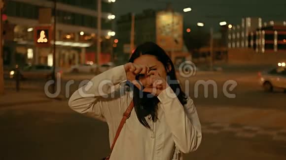 年轻的女人用手指做心脏符号把她漂亮的脸藏在夜城街的前边视频的预览图