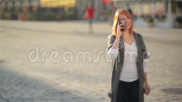 年轻的快乐兴奋的笑着女人在手机上说话红发女孩在城市的街头挥手视频的预览图