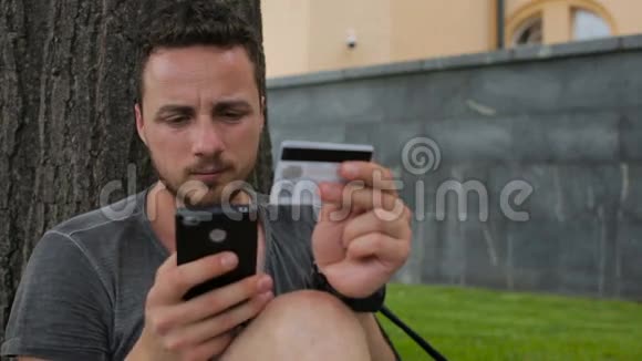 男子在公园里坐在树下通过互联网使用智能手机购物时使用信用卡视频的预览图