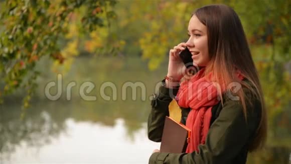 在湖边的秋园里年轻的女性在用手机说话美丽的女孩在笑公共公园的背景视频的预览图