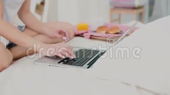 年轻女子在早餐时使用笔记本电脑坐在白色的床上女孩在电脑上打字和喝果汁的特写镜头视频的预览图